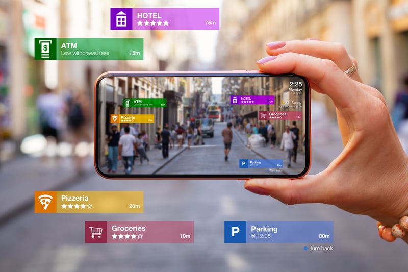 A person holding a smartphone displaying an augmented reality (AR) view of a busy city street. The AR interface overlays information about nearby locations such as hotels, ATMs, restaurants, and parking spots.