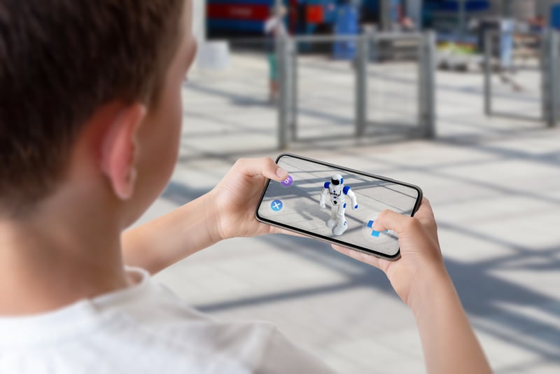 A young boy using a smartphone to interact with an augmented reality (AR) robot figure in a public space. The AR interface makes it appear as if the robot is standing in the real environment.