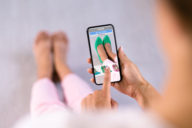 A woman using an augmented reality (AR) shopping app on her smartphone to virtually try on green high heels while sitting on the floor. The AR interface displays different shoe options.